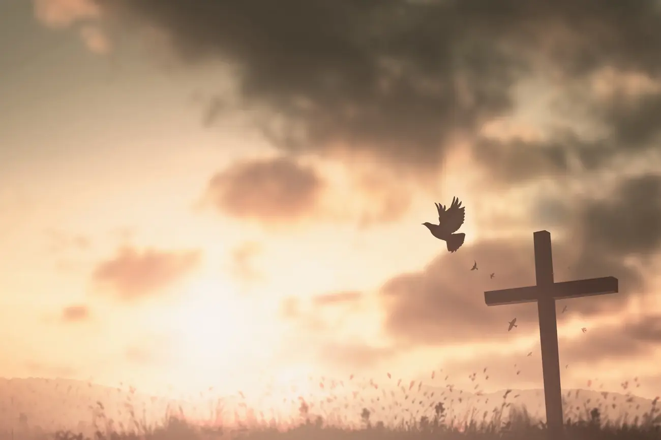 A dove flying toward a wooden cross at sunset